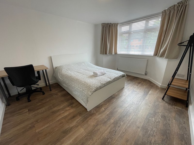 Bedroom with white bed, wooden flooring, desk chair, and large window with beige curtains