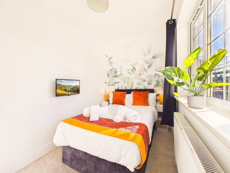 Bright bedroom with double bed, orange bedding, potted plant by window, and framed wall art