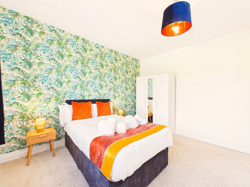 Bright bedroom with patterned green wallpaper, double bed with colorful bedding, wooden side table, and blue pendant light