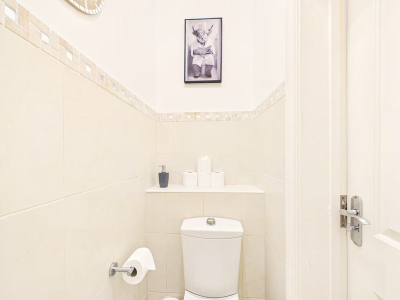 Small bathroom with toilet, wall-mounted shelf, framed artwork, and cream tile walls