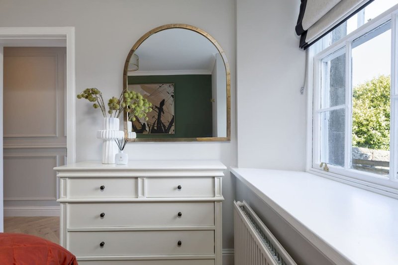 Bright bedroom with white dresser, round mirror, flowers in vase, and large window overlooking garden