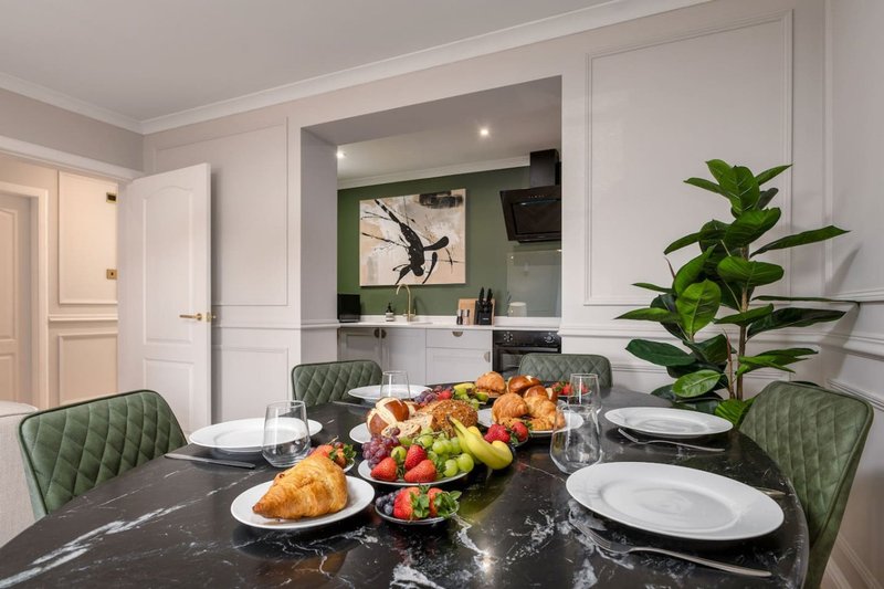 Modern dining room with black marble table, green quilted chairs, and open-plan kitchen with white cabinetry