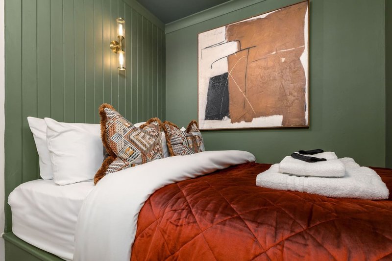 Bedroom with green panelled walls, double bed with rust-red quilt, patterned cushions, and framed abstract artwork