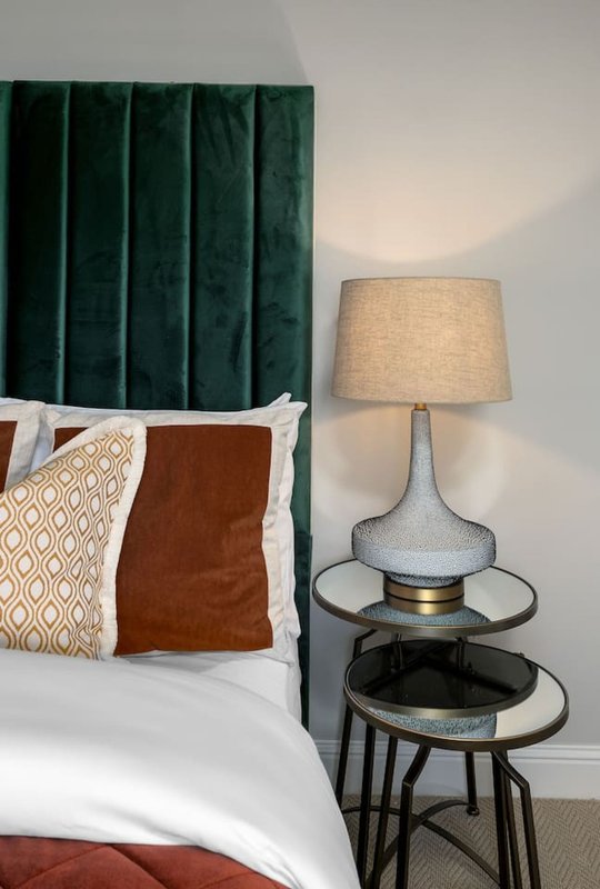 Bedroom with green velvet headboard, white sheets, decorative pillows, bedside lamp and side table