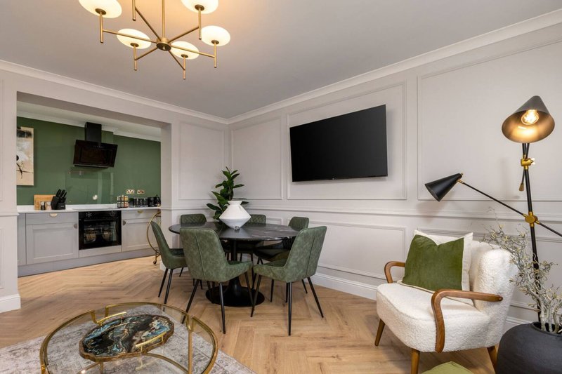 Open-plan living and kitchen space with dining table, green chairs, white armchair, and mounted TV