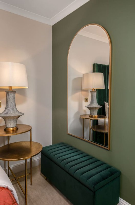 Modern bedroom corner with green accent wall, arched mirror, green upholstered bench, and brass table lamp