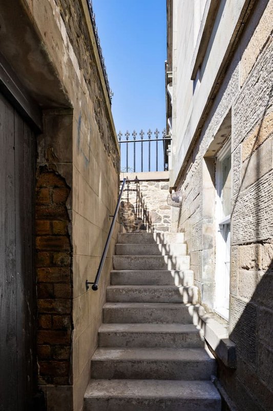 Narrow stone stairway between historic buildings leading to metal railing and clear sky