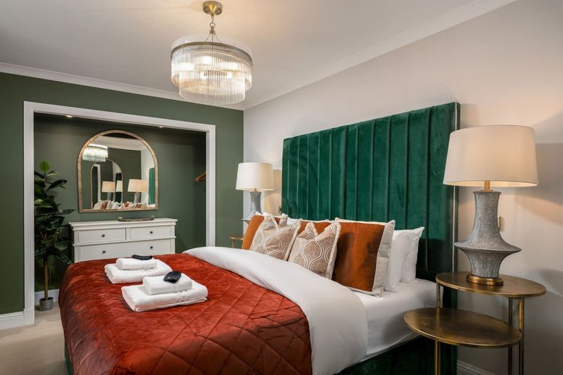Bedroom with green velvet headboard, white bedding, red quilted throw, and ensuite bathroom visible