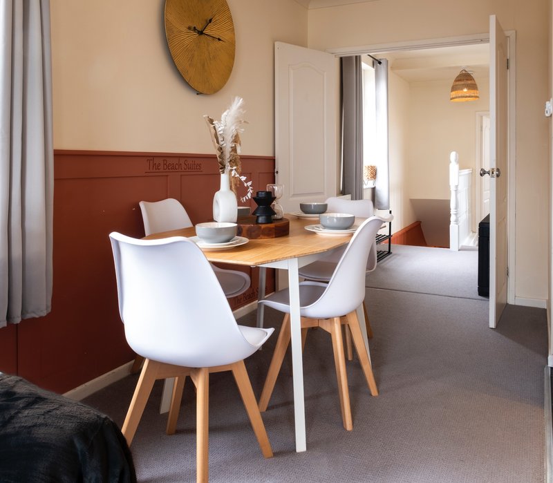 Modern dining area with wooden table, white chairs, wall clock and open doorway to bedroom