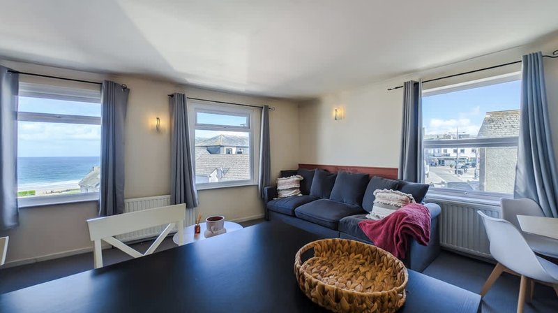 Bright living room with blue sofa, dining table, multiple windows overlooking coastal scenery