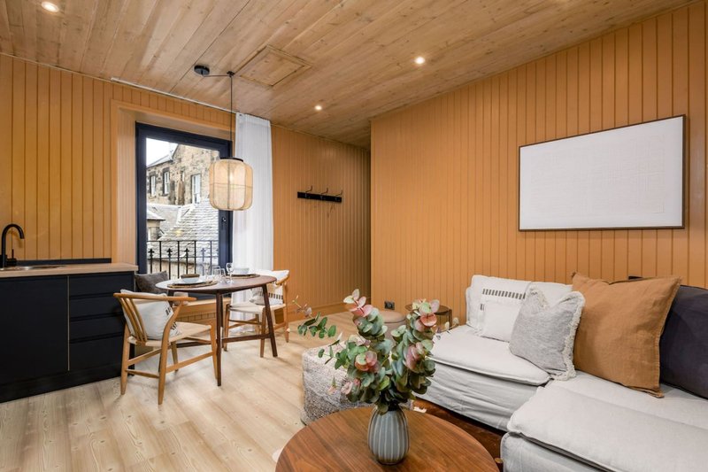 Warm wood-panelled studio with seating area, kitchenette, flowers in vase, and blank wall screen