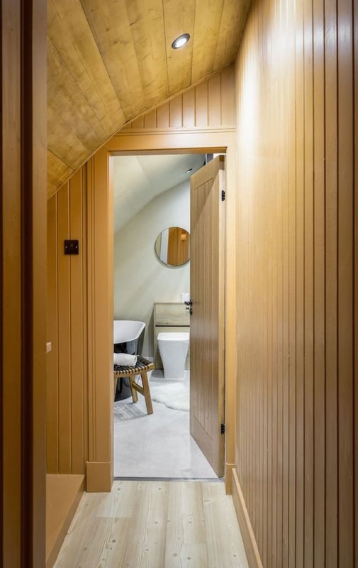 Wooden corridor with angled ceiling leading to bathroom with toilet and sink visible
