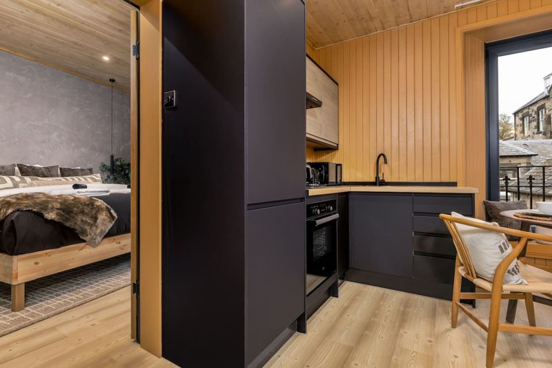 Modern studio apartment with black refrigerator, kitchen area, wooden furnishings and bedroom visible through doorway