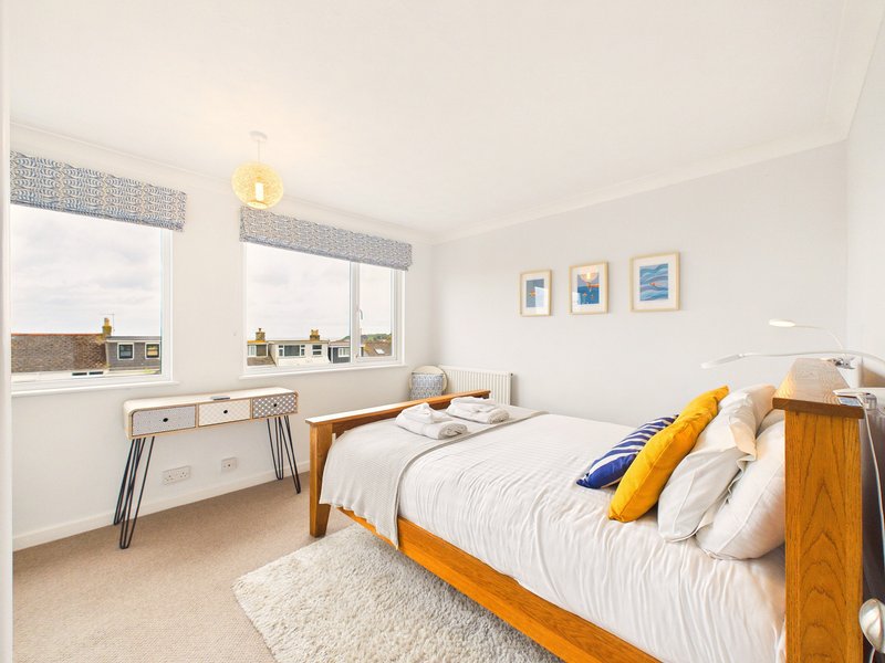 Bright bedroom with wooden bed, white bedding, yellow and blue cushions, desk by window