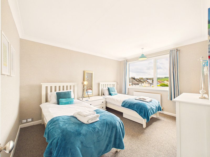 Bright bedroom with two single beds, blue bedding, white furniture and large windows overlooking coastal view