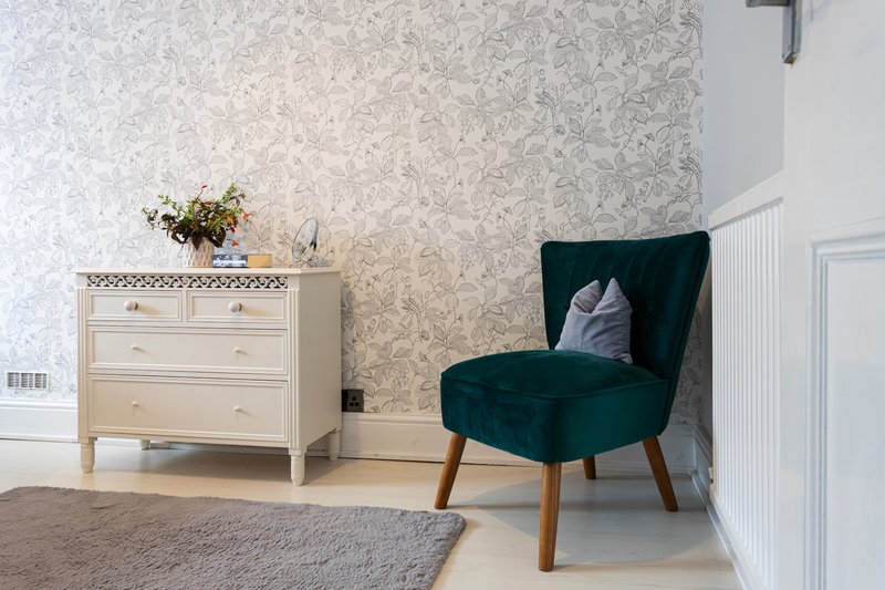 Bright bedroom with patterned wallpaper, wooden dresser, green armchair and wooden flooring