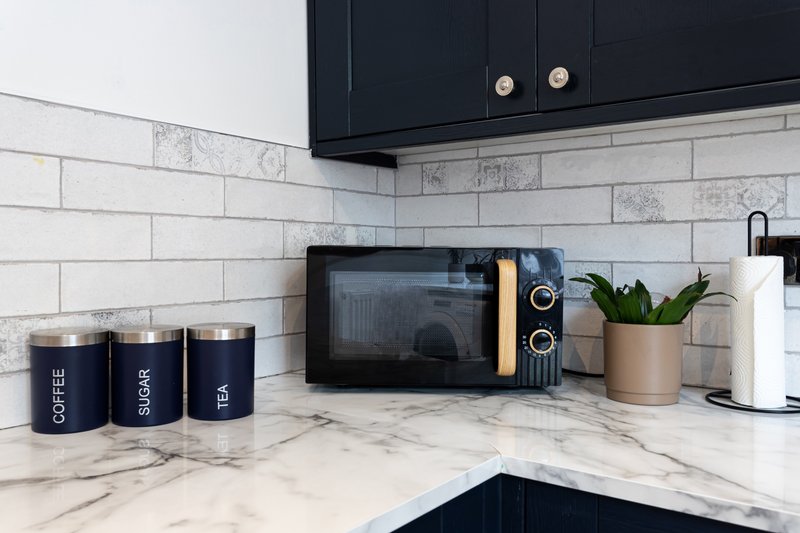 Modern kitchen corner with marble countertop, subway tile backsplash, dark cabinets, microwave and branded canisters