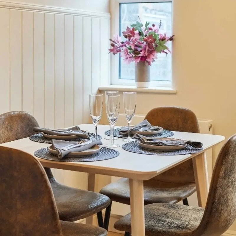Dining room with light wood table, brown upholstered chairs, place settings, and pink flowers in vase by window