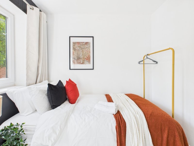 Bright bedroom with white bedding, red and black pillows, orange throw, and gold clothing rail