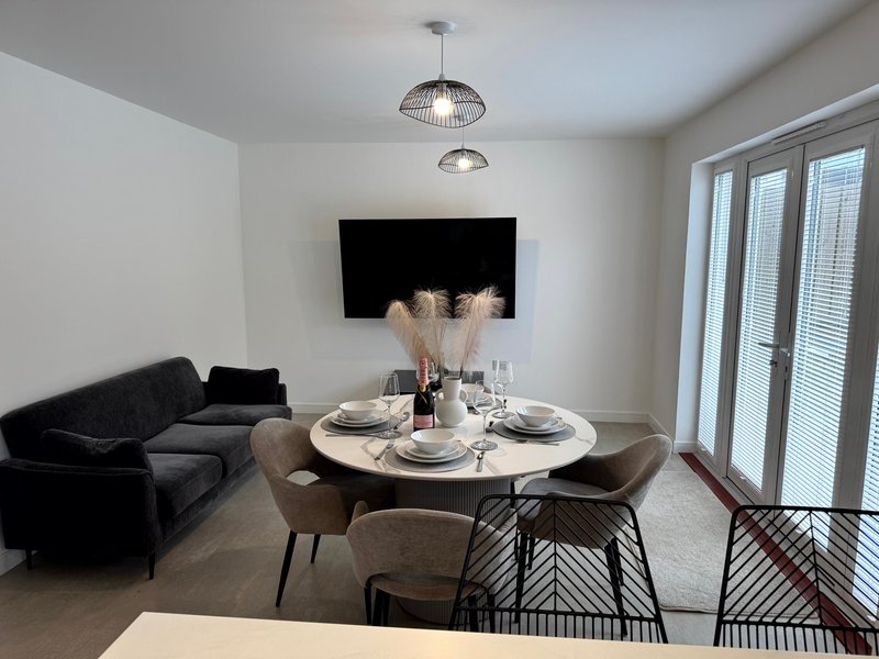 Modern living and dining area with black sofa, round white table, chairs, wall-mounted TV, and pendant lighting