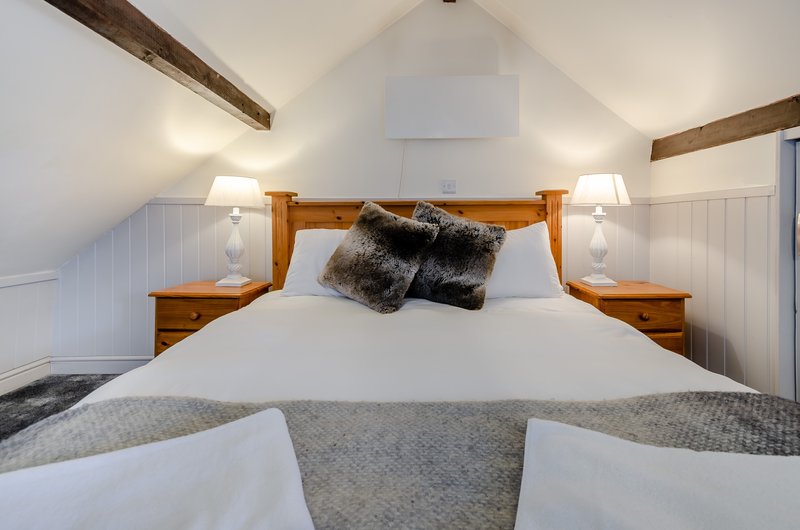 Bright attic bedroom with wooden bed frame, white bedding, dark fur pillows and exposed beams