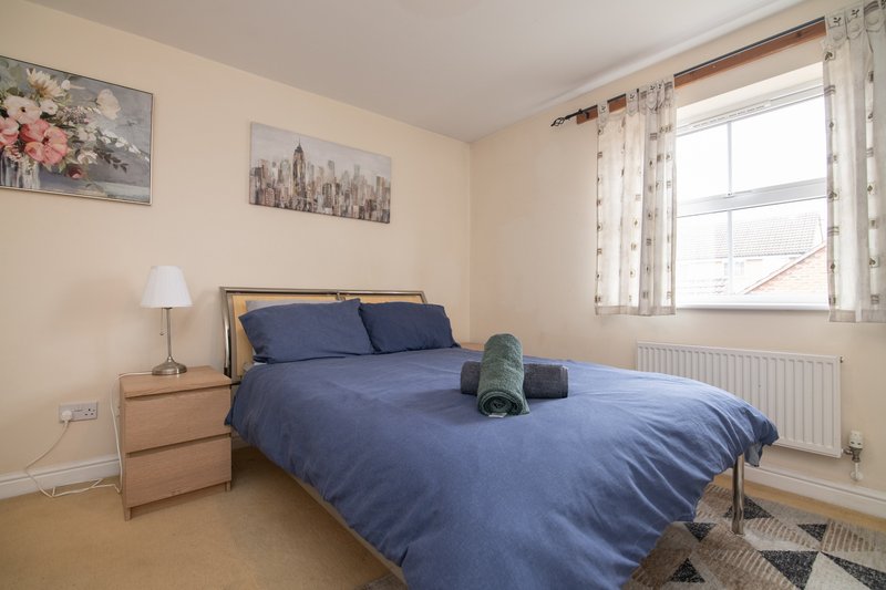 Bedroom with double bed, blue bedding, wooden nightstand, lamp, and window with curtains