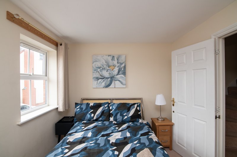 Bedroom with blue patterned bedding, wooden bedside table, window with curtains, and wall art