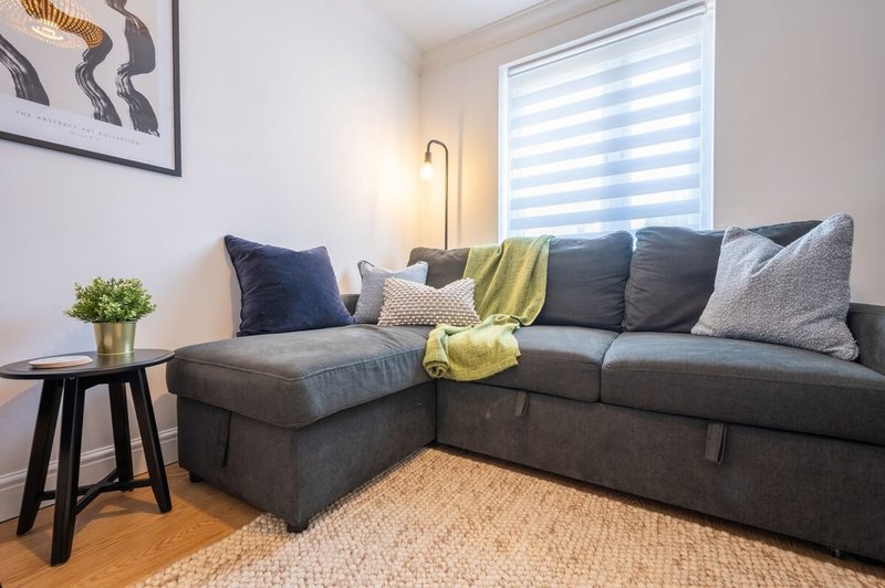 Modern living room with grey corner sofa, cushions, side table, and striped window blinds