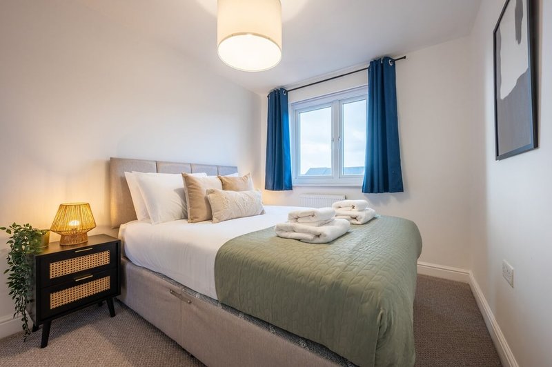 Bright bedroom with double bed, green bedspread, cushions, bedside lamp and blue curtains