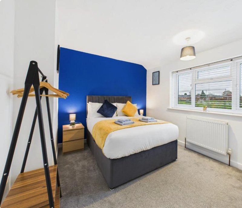 Modern bedroom with blue accent wall, double bed with yellow bedding, wooden coat stand, and large window