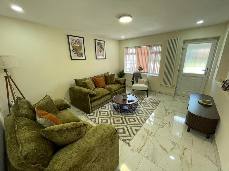 Bright living room with green upholstered sofa and armchair, patterned rug, framed wall art and glass doors