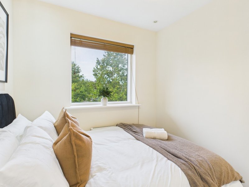 Bright bedroom with double bed, white bedding, tan cushions and window view of trees
