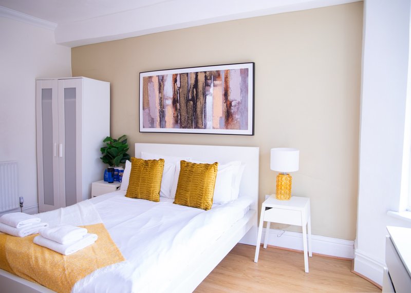 Bright bedroom with double bed, yellow cushions, white wardrobe and framed abstract artwork