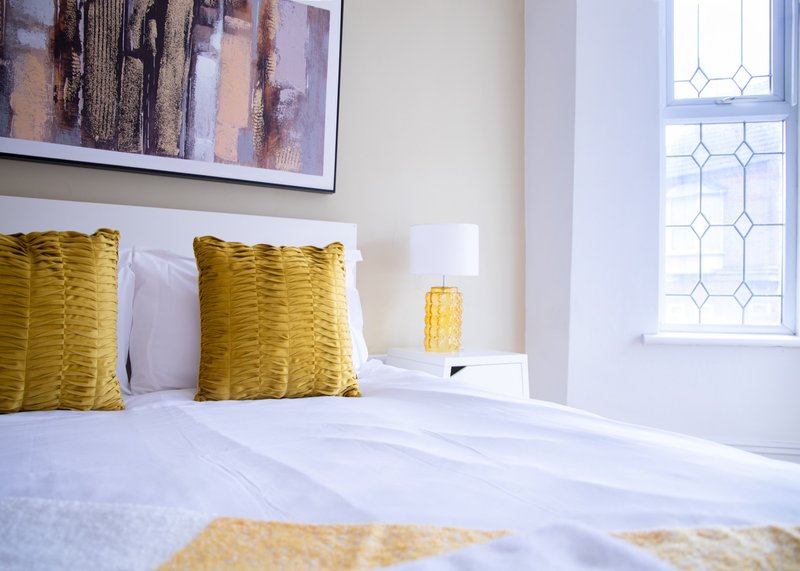 Bright bedroom with white bed, yellow cushions, framed artwork and decorative lamp