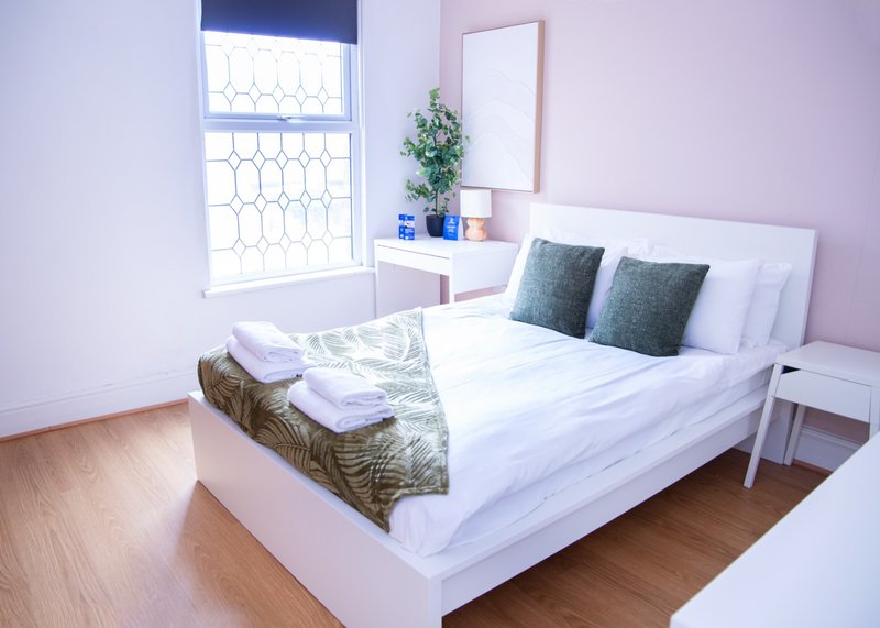 Bright bedroom with white double bed, pink walls, window with geometric blinds, and bedside table with plant