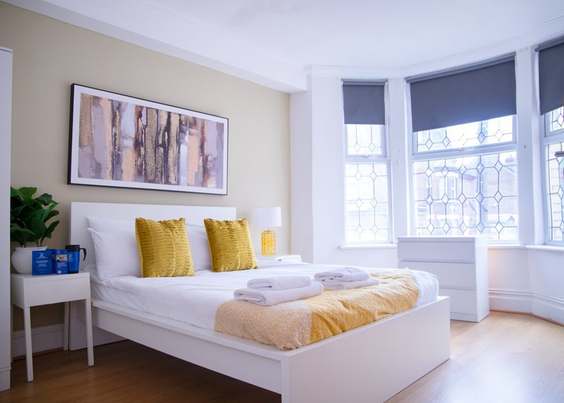 Bright bedroom with white bed, yellow pillows, framed artwork, side table and large windows