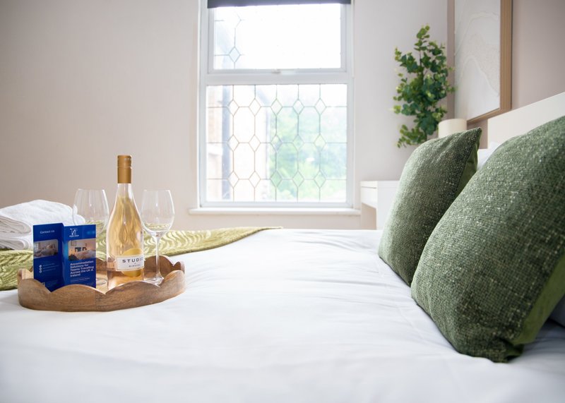 Bedroom with white bedding, green pillows, window with geometric panes, wine bottle and glassware on nightstand