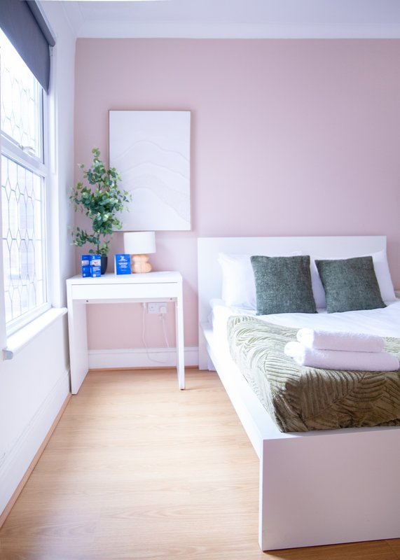 Pink bedroom with double bed, white desk, potted plant, and wooden flooring