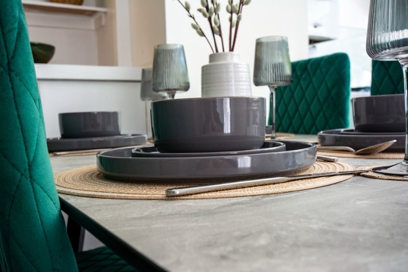 Modern dining table styled with grey dinnerware, green fabric chairs, and decorative vases with flowers