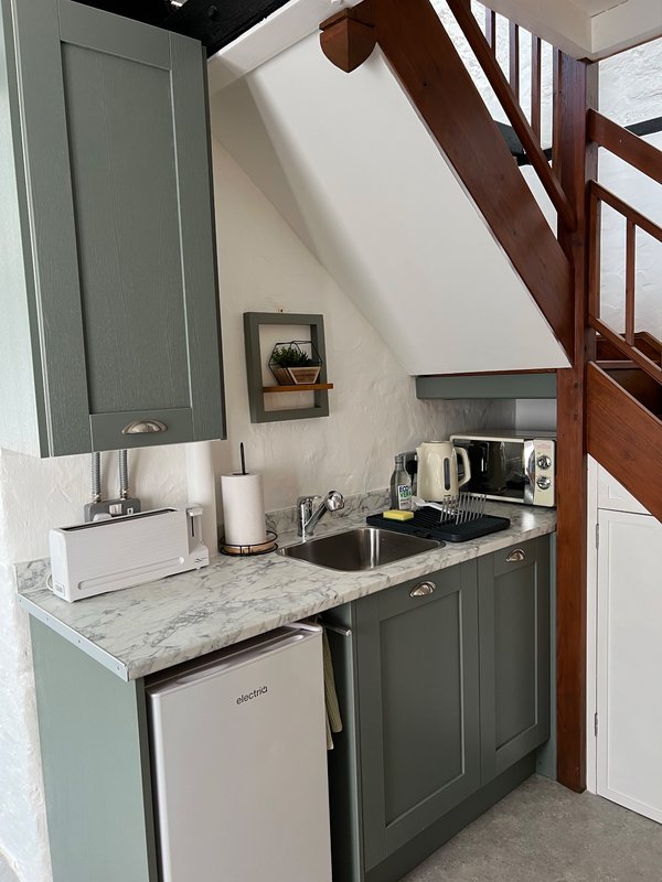 Compact kitchenette with grey cabinetry, marble countertop, sink and white appliances under wooden stairs