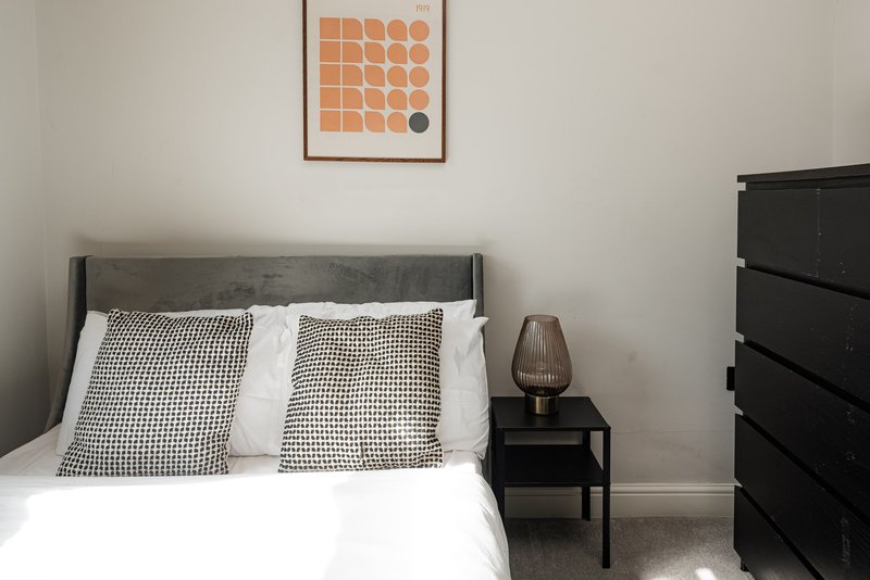 Bedroom with gray upholstered bed, patterned cushions, bedside table with lamp, and framed wall art