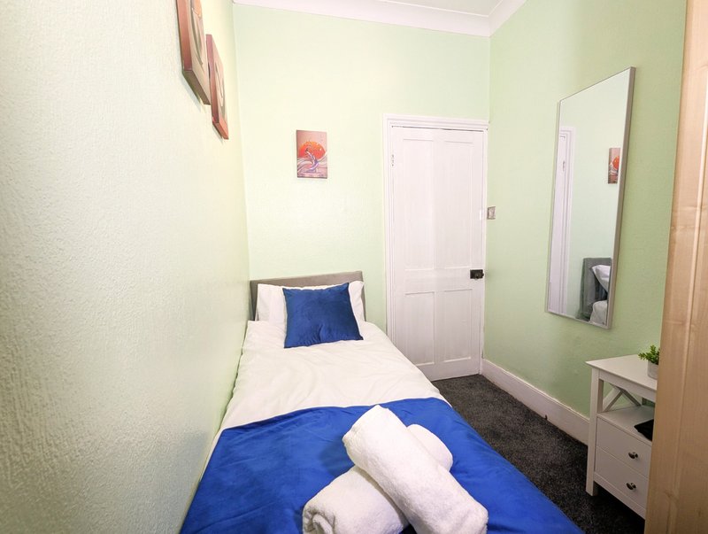 Small bedroom with single bed, blue bedding, white pillows, mirror and green accent wall