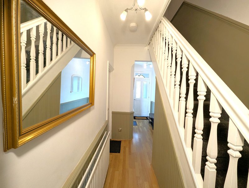 Narrow hallway with wooden flooring, white walls, staircase with black and white spindles, gold-framed mirror, and pendant light.