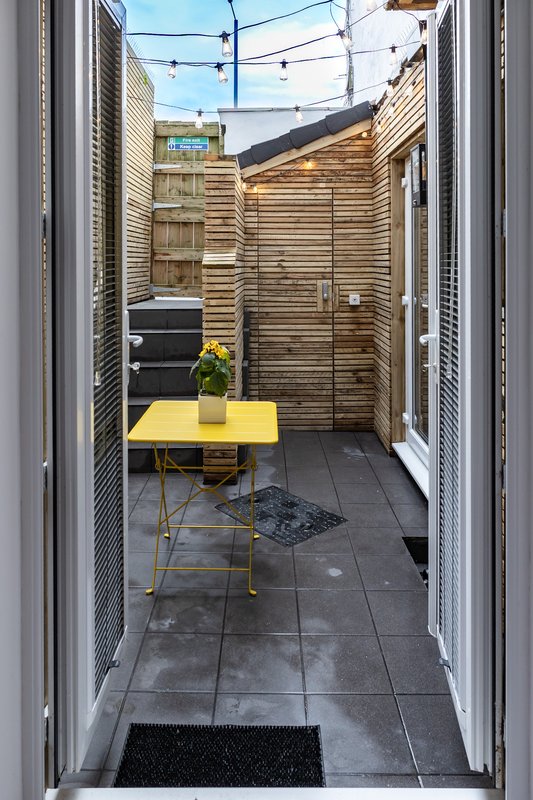 Narrow outdoor patio with wooden fence, yellow table, and potted plant overhead lighting