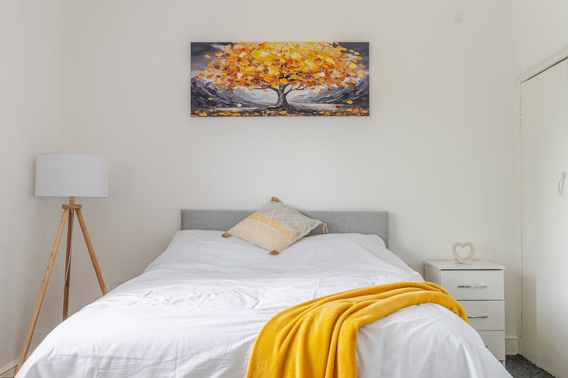 Bedroom with grey headboard, white bedding, yellow throw, wooden tripod lamp and wall art