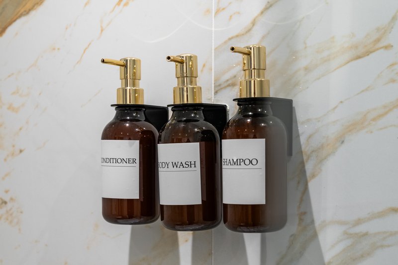 Three amber glass bathroom dispensers with gold pumps labeled conditioner, body wash, and shampoo