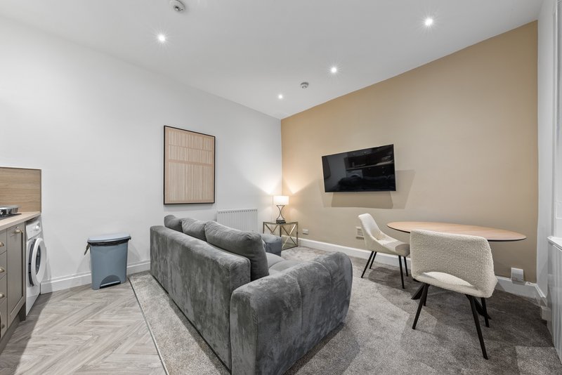 Modern living room with grey sofa, mounted TV, kitchenette, and neutral decor