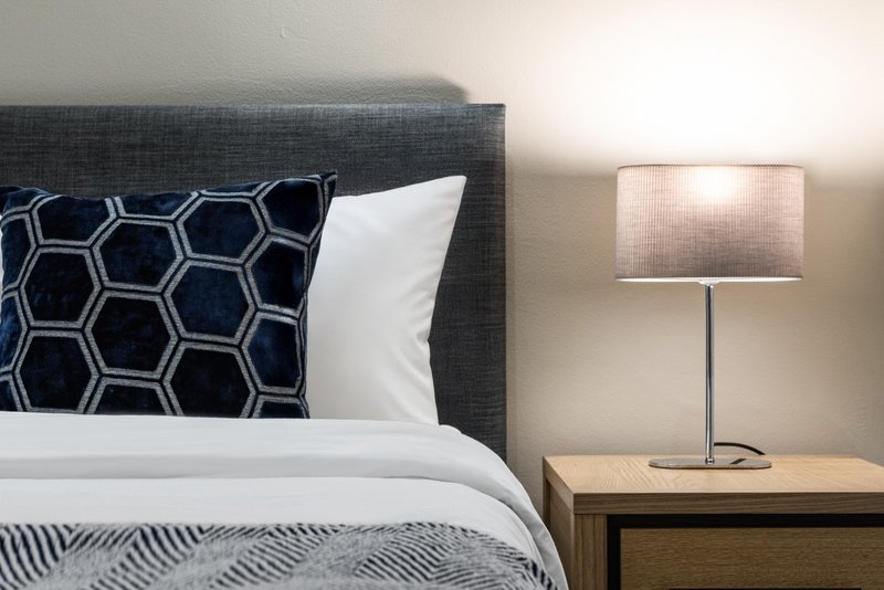 Modern bedroom with gray upholstered headboard, patterned cushion, white pillows and bedside lamp