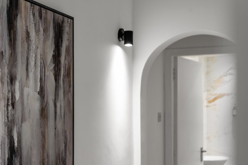 Modern white hallway with arched doorway, wall sconce lighting, and abstract artwork
