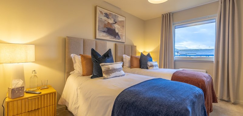 Bright bedroom with two double beds, bedside lamps, blue throw blanket and ocean view through large window